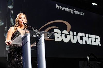 AVP Chantal Beaver standing on stage at gala  speaking into microphone with "presenting sponsor BOUCHIER" behind her.