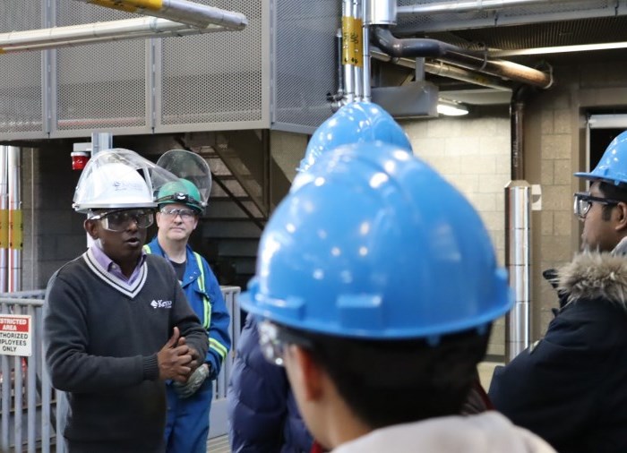 instructor with safety glasses and hardhat standing in front of a group of students with safety glasses and hardhats on in a power engineering lab instructing the students