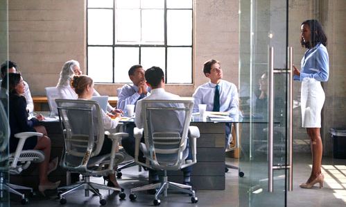 coworkers in a board room at a table listening to a woman standing talking