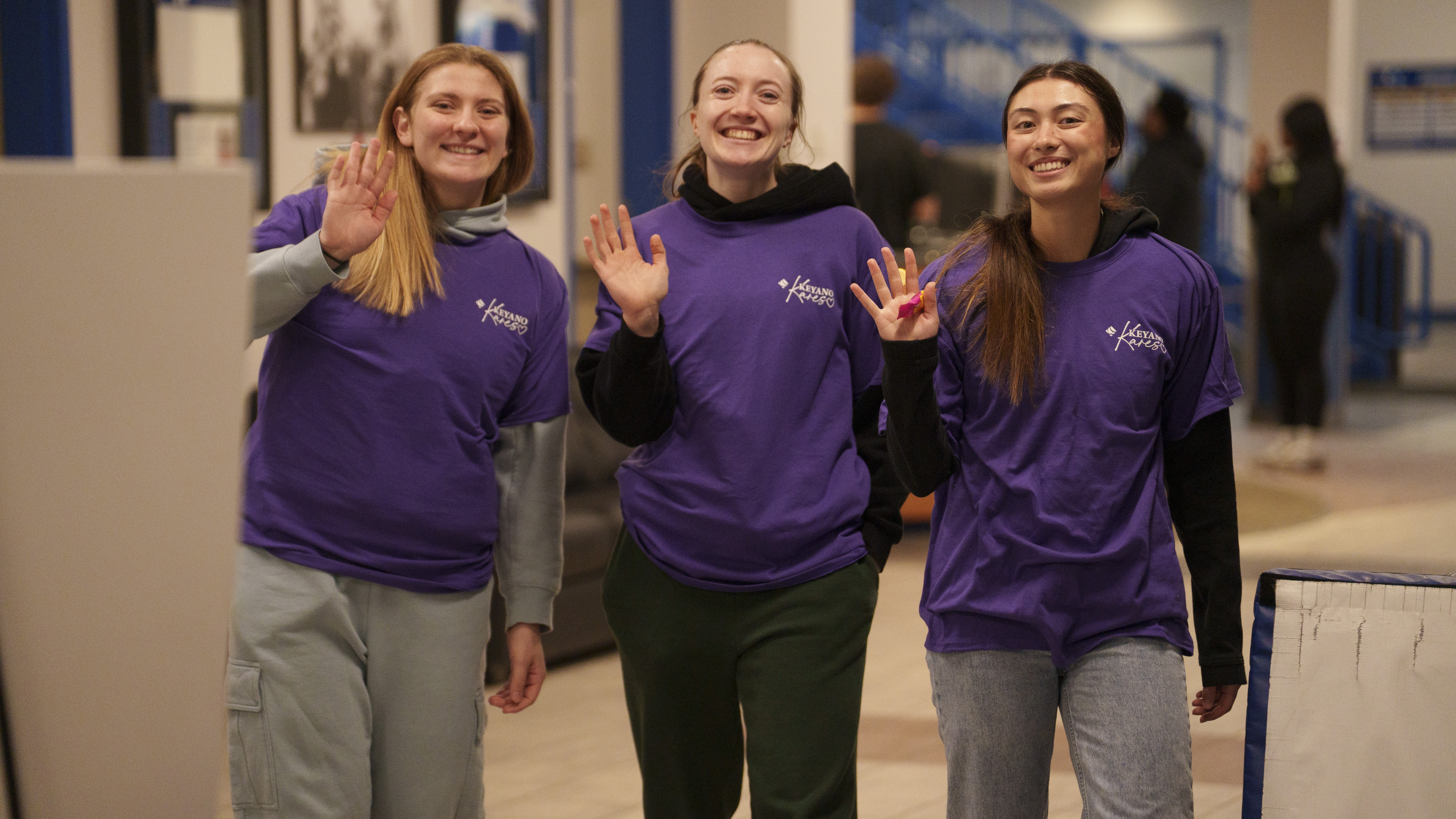 Three students waving their hands at the camera. They are wearing purple tshirts with Keyano Kares logo at the top right corner