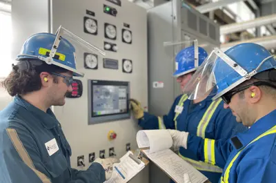 Three power engineering students wearing hard hats, face shields, and safety gear reviewing operational checklists beside an industrial control panel.
