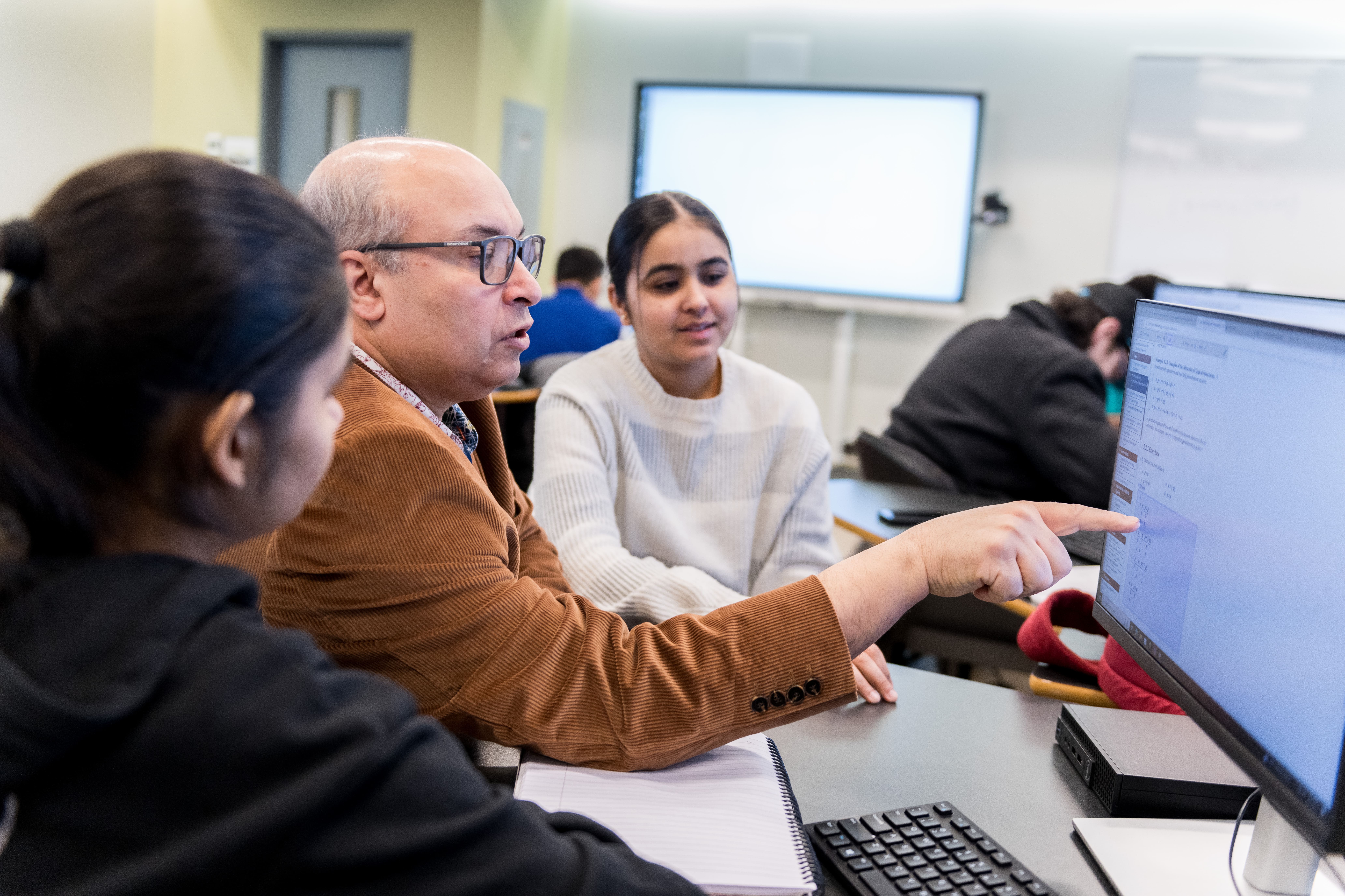 A professor pointing at a computer and 2 students are sitting beside him
