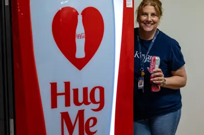 A staff standing next to a Hug Me machine with a can of coke in her hand