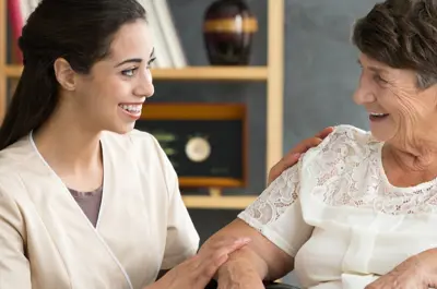 A health care aide smiles and talks with an older adult seated in a wheelchair.