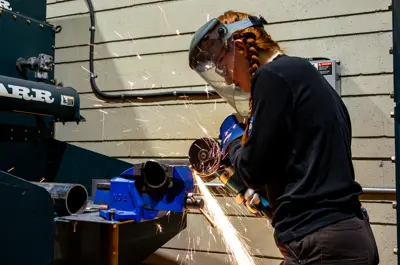 Student wearing protective equipment using a grinder on metal piping in a trades workshop, with sparks flying during metalwork.