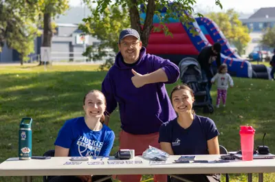 Three people smile at an outdoor community event while seated and standing behind an information table. In the background, families enjoy activities near an inflatable play structure in a park setting.