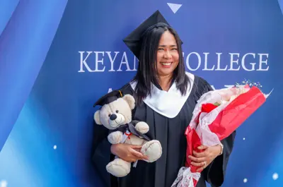 A smiling graduate holds a teddy bear and bouquet of flowers in front of a Keyano College backdrop.