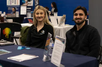 A woman and a man sitting side by side behind a table that has tickets, forms, and a bunch of papers smiling for the camera