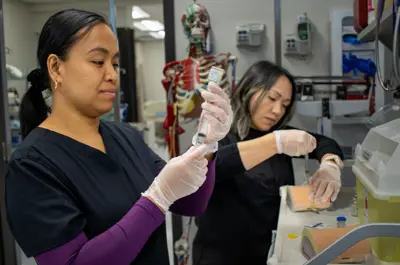Two healthcare workers wearing gloves prepare and handle syringes at a clinical workstation, with medical equipment and an anatomical model visible in the background.