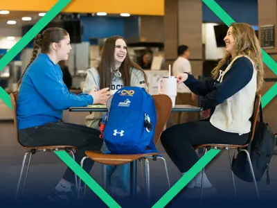 Three students sit around a table in a campus common area, smiling and chatting over coffee. A backpack rests beside them