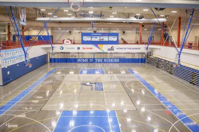 Wide view of the Keyano Huskies gymnasium at the Syncrude Sport and Wellness Centre. The indoor facility features a full-size basketball court, multiple hoops, spectator seating, and prominent “Home of the Huskies” branding throughout the space.