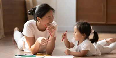 Educational pastime develop creativity skill in kid concept. Asian mother her small daughter lying on warm wooden floor in sunny cozy living room, mom teach girl paint use album and colourful pencils
