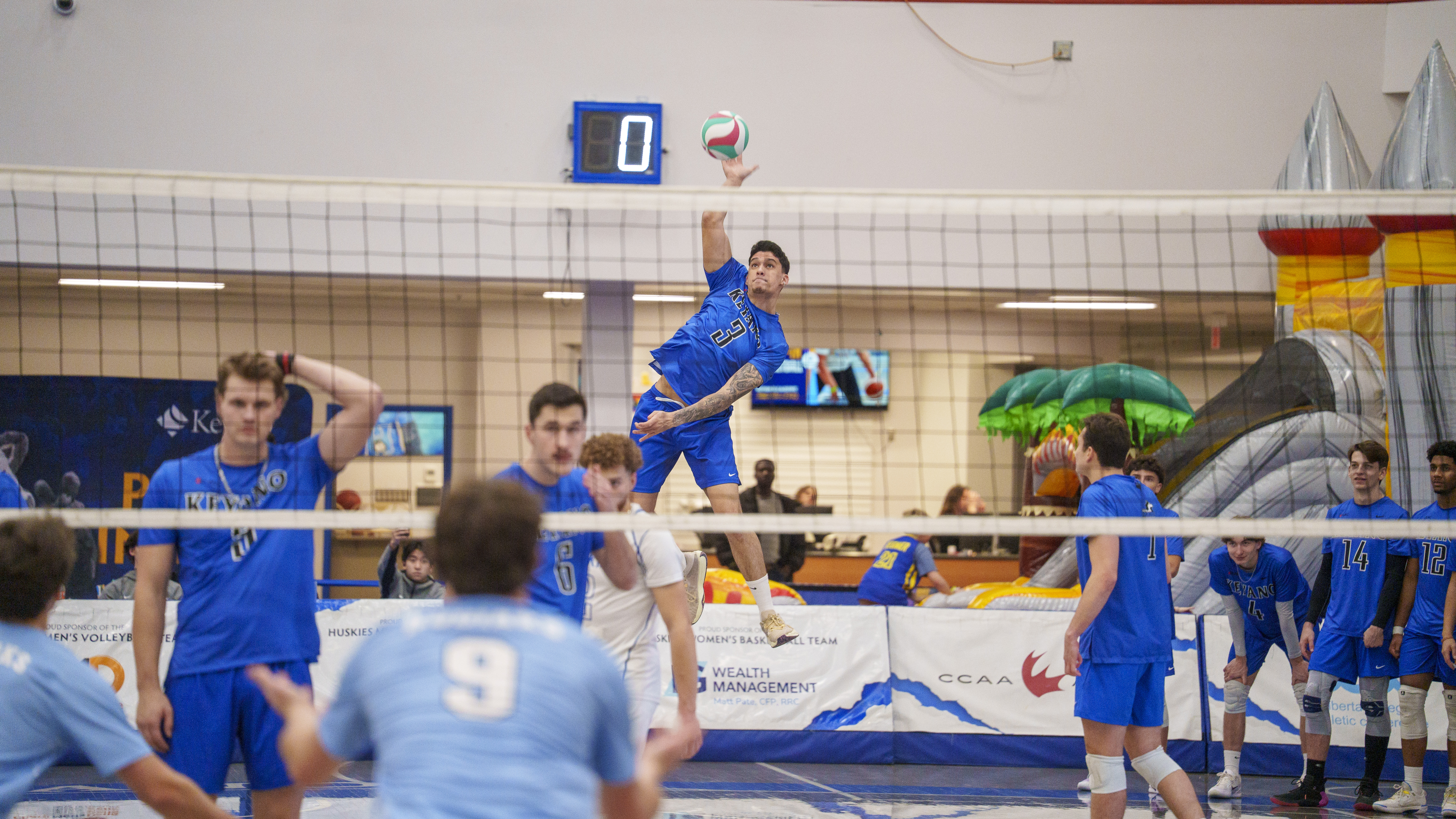 Indoor volleyball match in a gymnasium, with a player in a blue jersey (#21) airborne mid-spike over the net, while the opposing team, also in blue uniforms, prepares to block or receive the ball. A scoreboard and inflatable structure are visible in the background