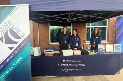 Three staff members stand behind a Keyano College Alumni Association information booth at an outdoor event.