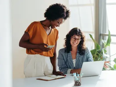 Happy professional women collaborating with enthusiasm in a modern office setting. Two female entrepreneurs using a laptop for a productive business meeting in a corporate workplace.