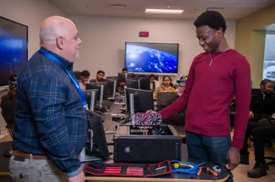 An instructor speaks with a student who is installing a graphics card into a desktop computer during a computer hardware lab, while other students work at computer stations in the background.