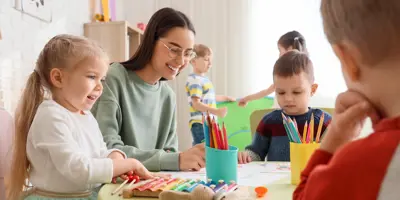 Cute little children with nursery teacher drawing at table in kindergarten