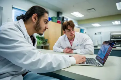 Two students wearing lab coats collaborate on a laptop in a science laboratory, reviewing data and discussing their work during a lab session.