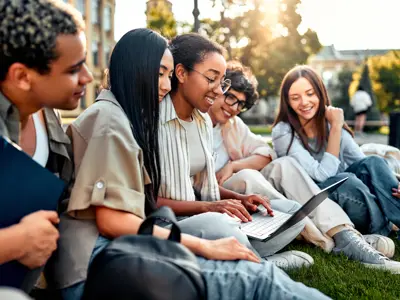 Group of happy diverse students sitting on grass and studying with laptop on university campus. Student life, high school. Motivation and learning.