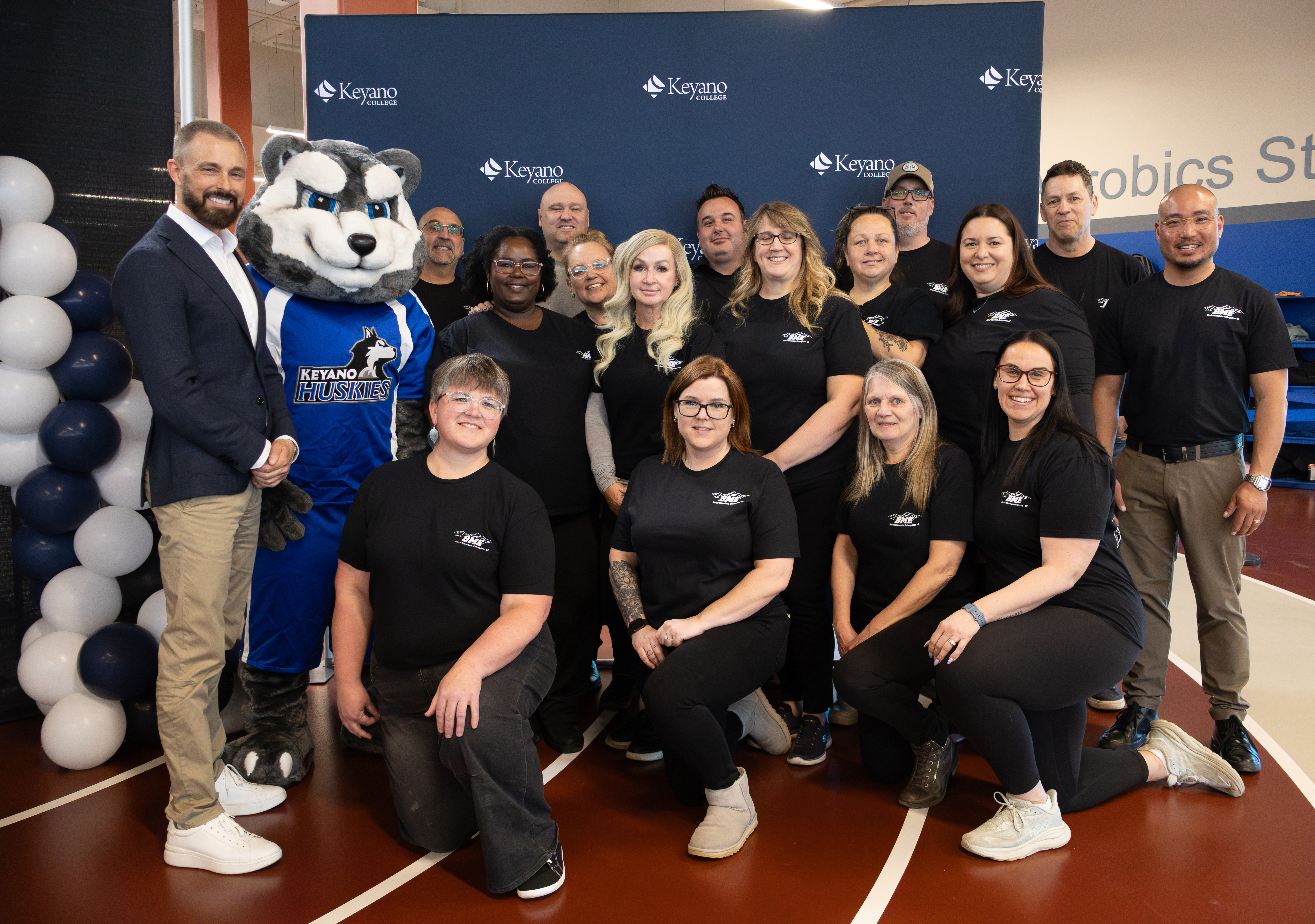 Group photo of Keyano College President & CEO Dr. Scott MacPherson, BME staff standing with the Huskies mascot in front of a branded backdrop at an indoor event.