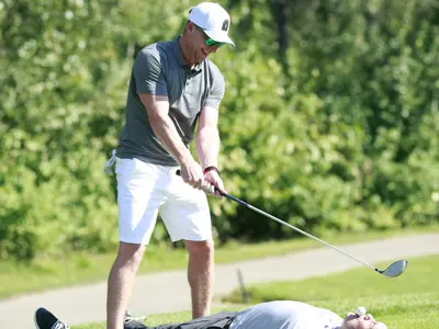 A man aims at a golf ball held in another man’s mouth as he lies on the grass.