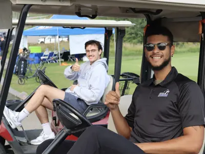 A man in sunglasses strikes a playful pose in front while another man gives a thumbs up sitting on a golf cart behind him.