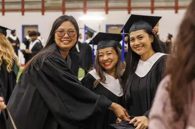 Three graduates wearing caps and gowns smile together at a graduation ceremony.