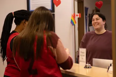 A smiling staff member assists two visitors at a reception desk decorated with Canadian and Pride flags.
