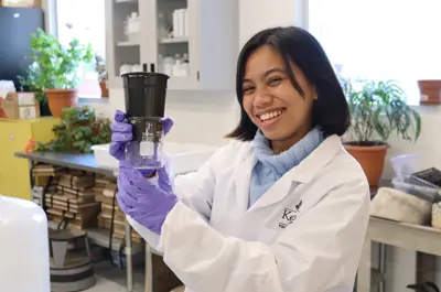 A smiling student wearing a lab coat and purple gloves holds a beaker and plant pot in a laboratory.