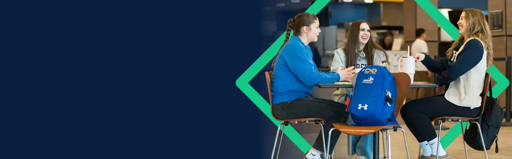 Three students sit around a table in a campus common area, smiling and chatting over coffee. A backpack rests beside them 