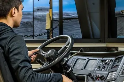 Student operating a haul truck simulator with hands on the steering wheel and controls in a mining training environment.