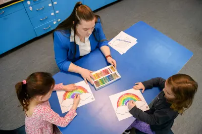 An early childhood educator helps two children colour rainbow pictures at a classroom table.