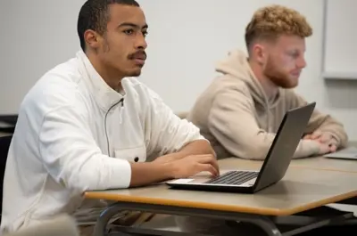 Two students seated in a classroom, working on laptops and listening during a lesson.