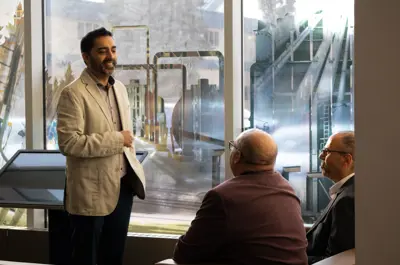 A guy in a white vest standing talking to 2 guys who are sitting down